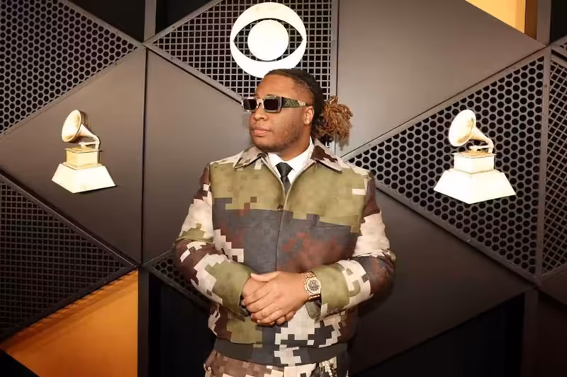 Tay Keith standing on the red carpet at the Grammy Awards wearing sunglasses and a dark jacket.